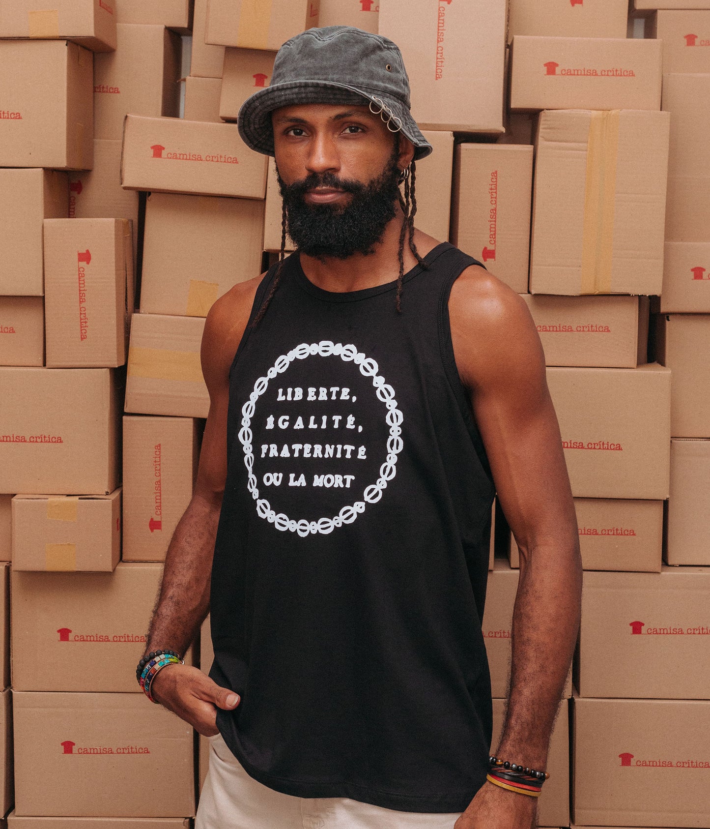 Homem vestindo Camiseta Regata. Estampa: Insígnia da Revolução Francesa. texto com os dizeres em francês: Liberte, Egalite, Fraternite ou La Mort. Truz: Liberdade, Igualdade, Fraternidade ou a Morte. Fios transados circulam o texto.