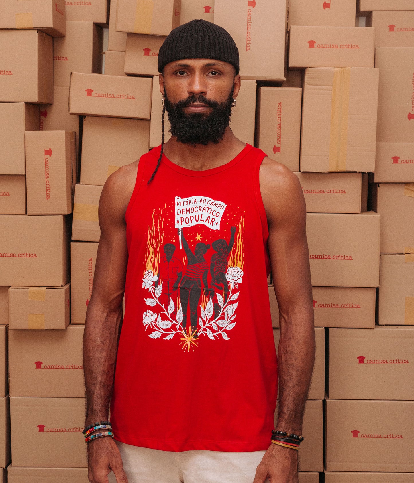 Homem vestindo Camiseta Regata. Estampa: Três pessoas caminham como se fosse uma passeata, ao centro uma mulher leva uma bandeira com o texto: Vitória ao campo democrático popular. A imagem é emoldurada com flores e chamas embaixo e ao lado.